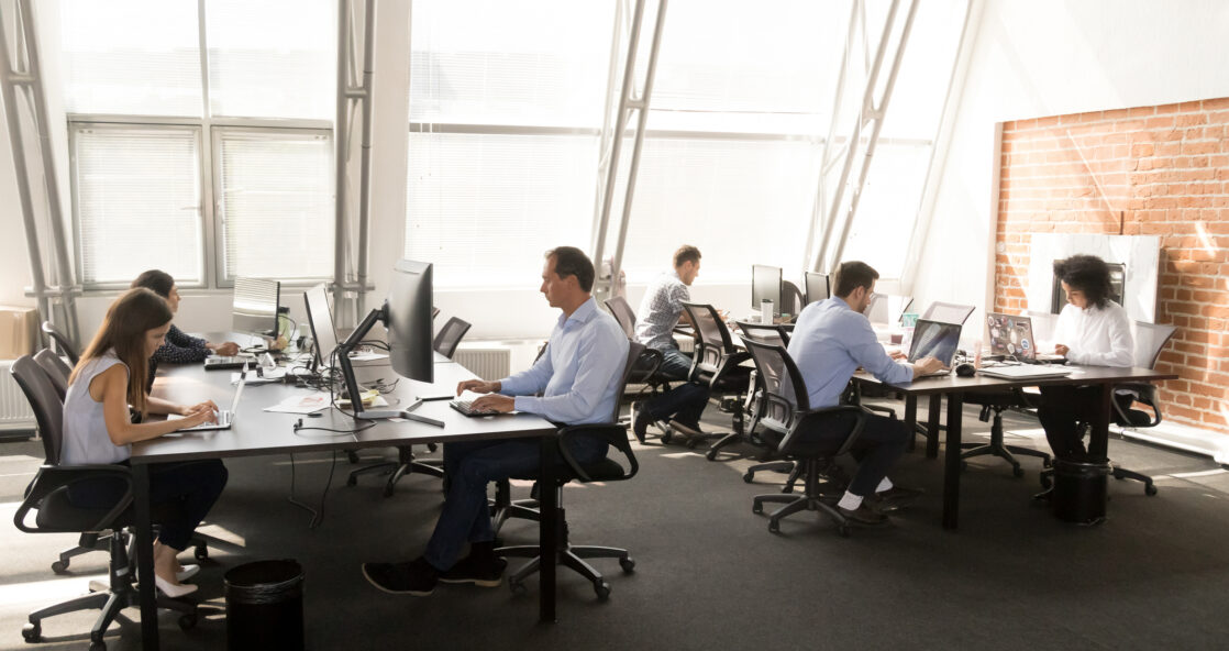Diverse multiracial workers sitting at desk working in coworking space. Busy six businesspeople coworkers using computers typing chatting solving business matters in modern light contemporary office